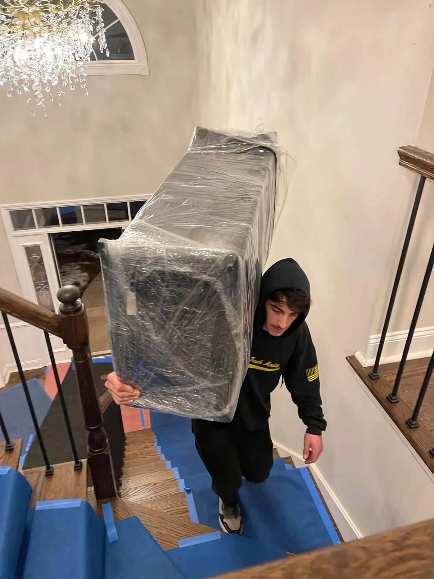 Yellow Truck Moving mover carrying a shrink-wrapped chair up a taped-off staircase in a luxury home